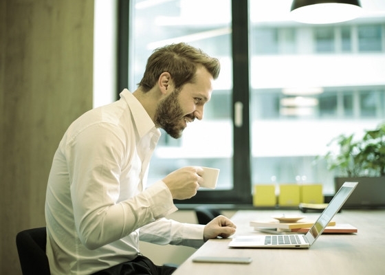 a happy man in sitting in a coffee shop front of a laptop holding a cup