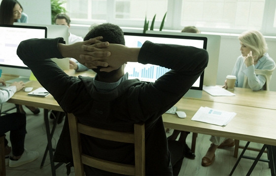 a man with his hands behind his head in front of a computer in a conference room with others